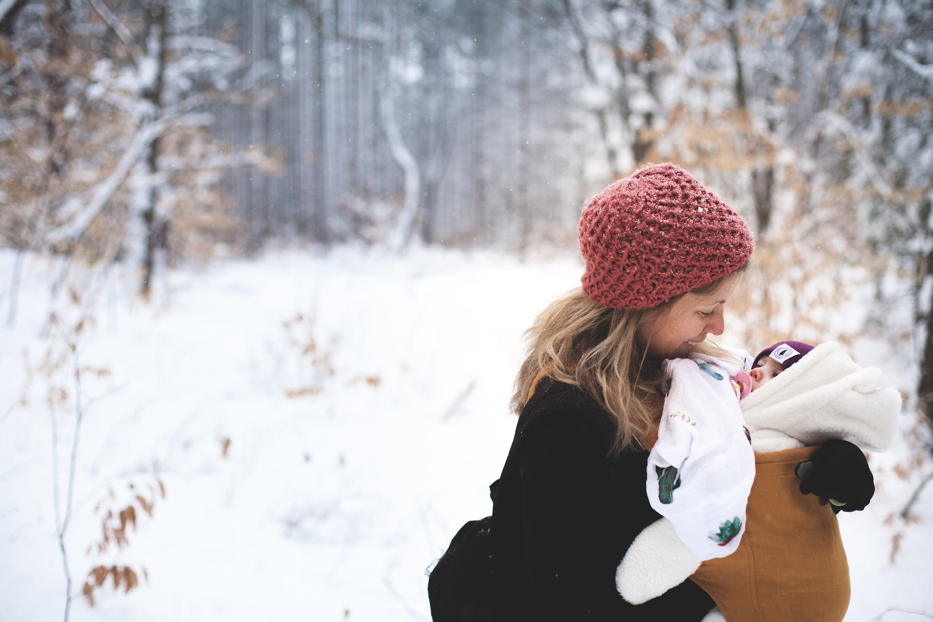 A mom looks at her baby that lays on her in a baby carrier out in a snowy forest.