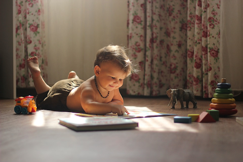 Baby laying on floor reading picture book while surrounded by toys.