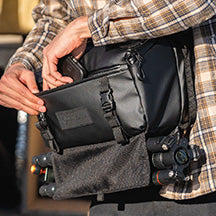 A person wears the Roam 9 Liter Sling on the front of their body, sliding their phone into the front zipper pocket. A camera tripod is attached to the bottom of the bag.