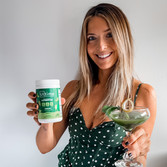 Woman holding martini glass with green drink in one hand and a tall canister of Ultima Replenisher Appletini flavor electrolytes in the other hand