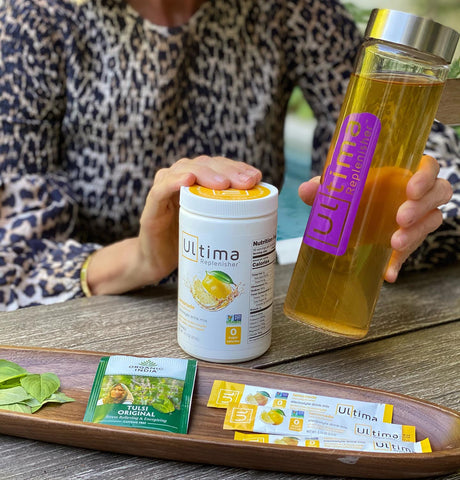 Woman holding Wellness Tonic made with Ultima Replenisher and a 90 serving canister of Lemonade electrolyte hydration powder