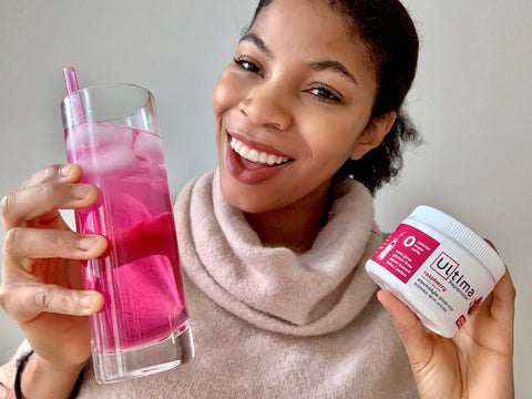 Woman holding a container of Ultima Replenisher Raspberry flavored electrolyte powder and Ultima Replenisher raspberry in a glass