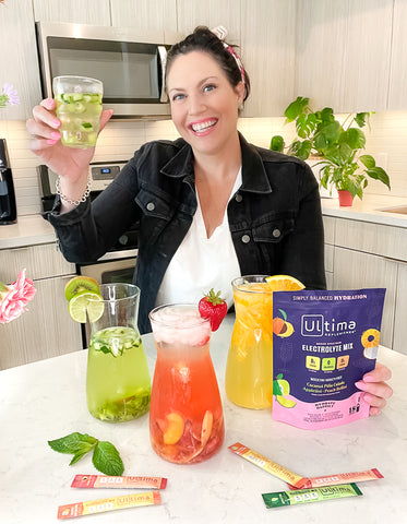 Woman holding up a glass of Ultima Replenisher with pitchers and stickpacks  of different flavors