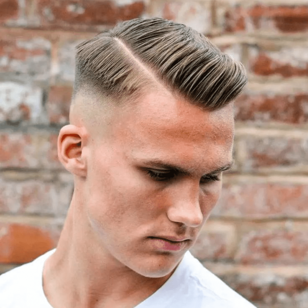Close-up of a young man with a sharp casual side part hairstyle, featuring clean lines and a sleek finish, styled with pomade against a brick wall background