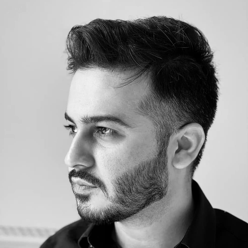 A black-and-white profile portrait of a man with a neatly groomed short boxed beard. His beard is full but trimmed short, following clean, well-defined lines along his cheeks and jawline. This short beard style enhances his sharp facial features and provides a balanced, sophisticated look. The man has short, neatly styled hair, with the top slightly longer and brushed back for a polished appearance. He is wearing a dark shirt, adding to his refined and professional demeanor. The background is plain and uncluttered, ensuring that the focus remains on his face and beard. The lighting highlights the texture and grooming precision of his beard, giving the portrait a crisp and clear look.