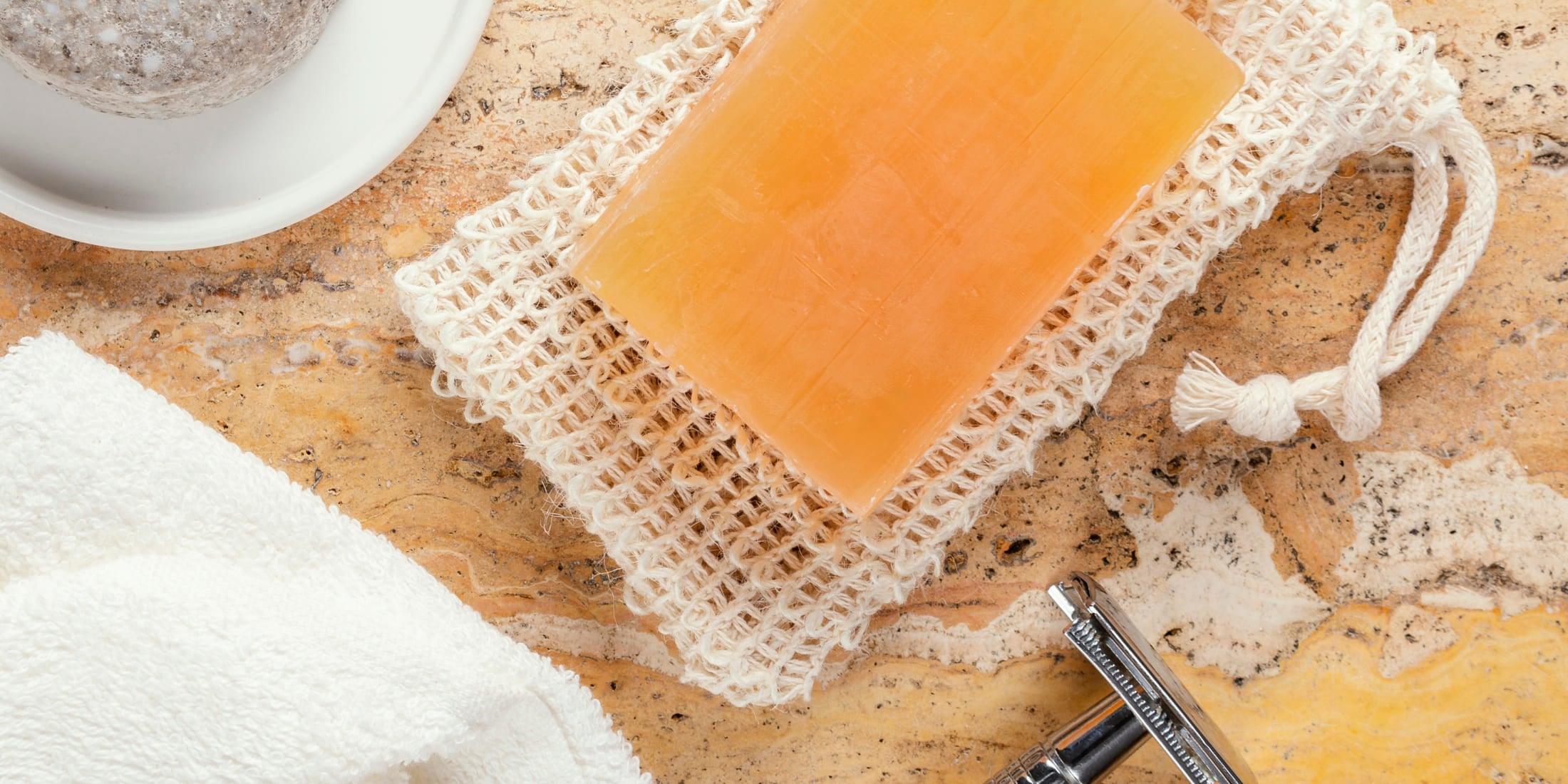 Bar of shaving soap on a natural exfoliating pad next to a classic safety razor and a soft white towel on a marble countertop. The image highlights traditional wet shaving essentials, showcasing the rich texture and natural ingredients of the shaving soap.