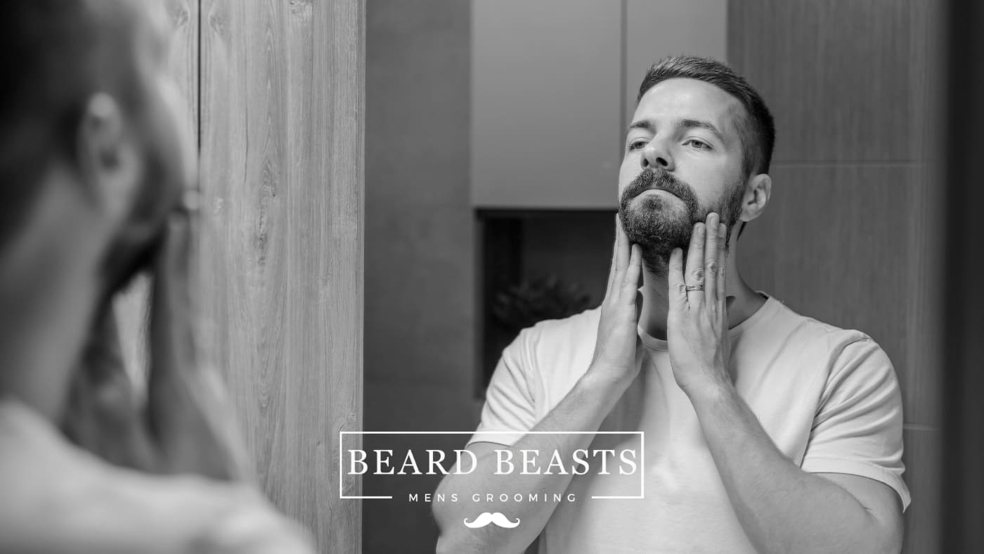 A man thoughtfully touches his beard in front of a bathroom mirror, a moment of self care for men
