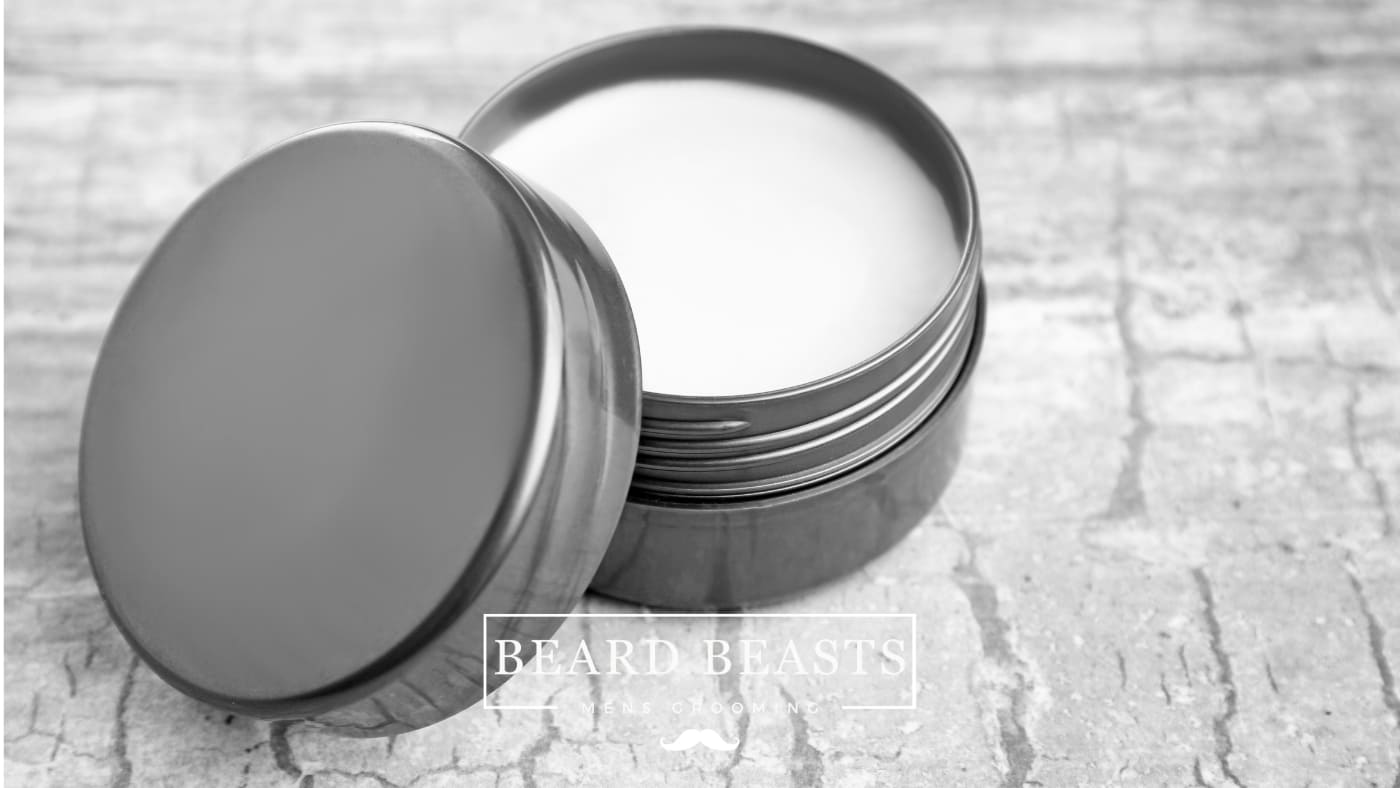 Black and white image of an open tin of pomade on a textured surface with the lid placed beside it. This image hints at the theme of Oil Based vs Water Based Pomade for hair styling products.