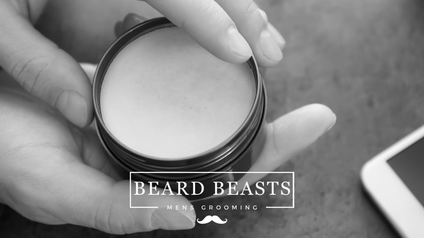 Open tin of hair styling product held in hands with 'BEARD BEASTS - MEN'S GROOMING' logo, symbolizing the choice between hair clay vs wax for men's styling needs, alongside a smartphone on a muted background.