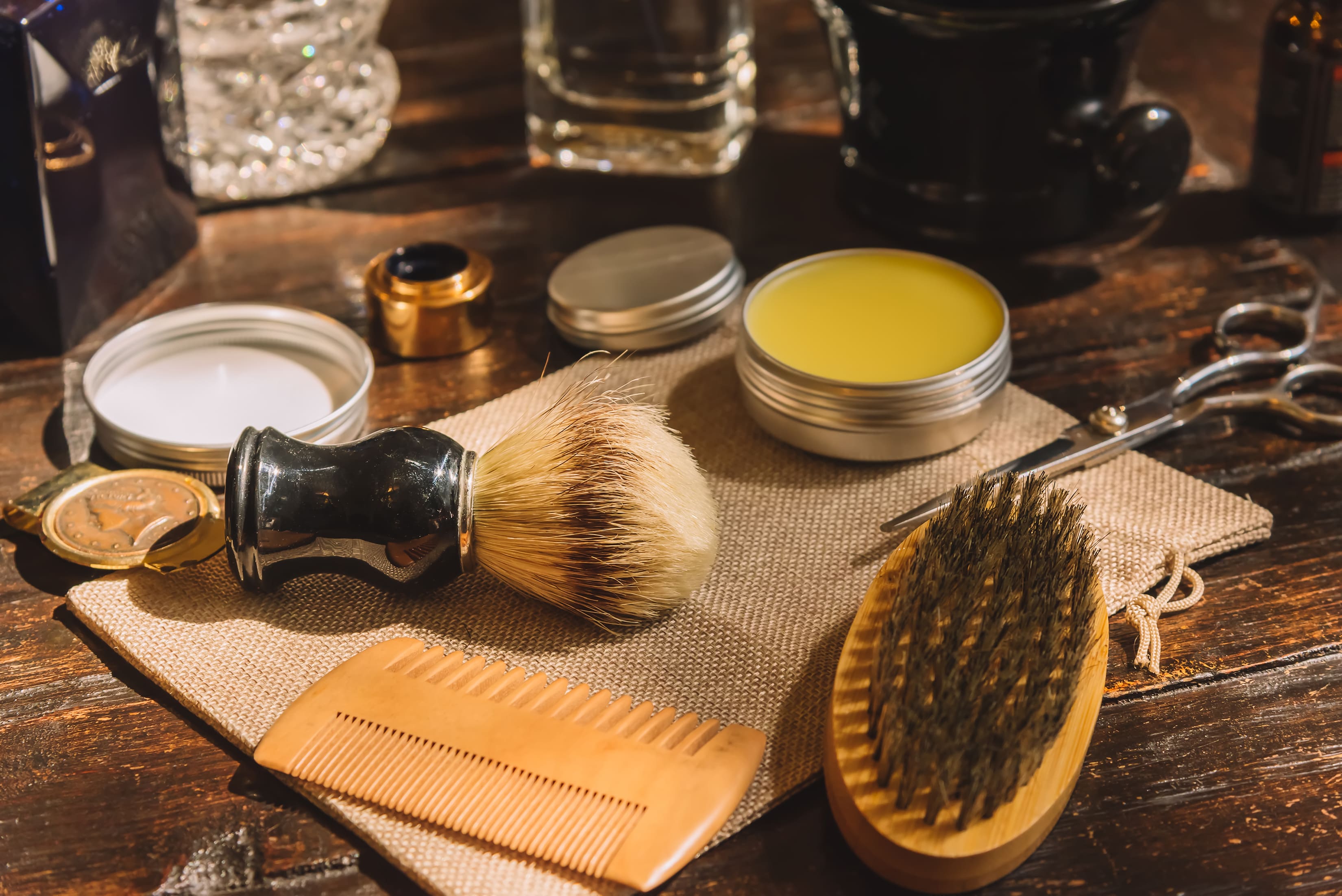 A rustic wooden table displays various beard grooming tools, including a shaving brush, scissors, combs, and tins of beard balm and wax. These essentials are artfully arranged to showcase the key products needed to tame unruly beard hair and prevent stray hairs from sticking out, ensuring a well-groomed and polished beard.