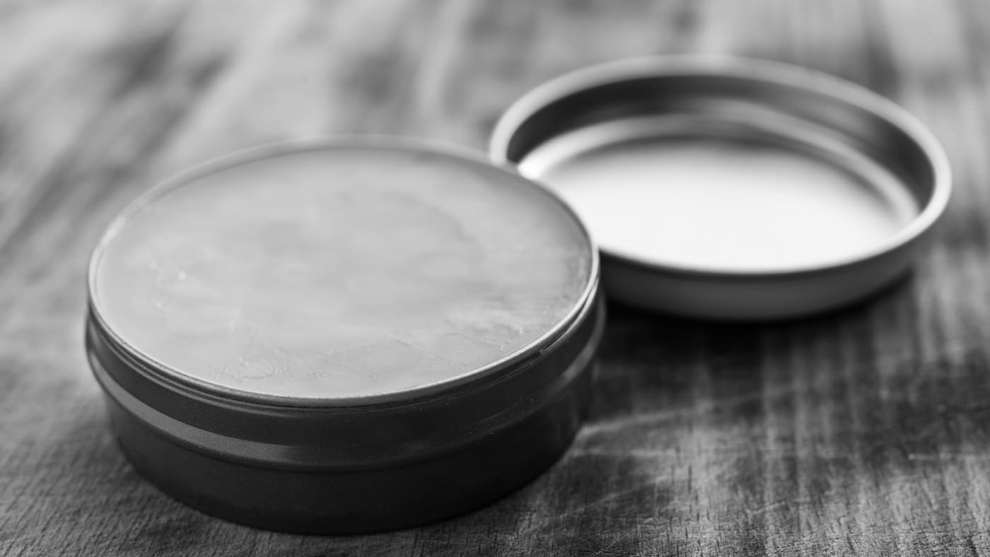 Mustache wax vs beard balm comparison, with open tins on wooden background, in monochrome for focused product texture and usage details.