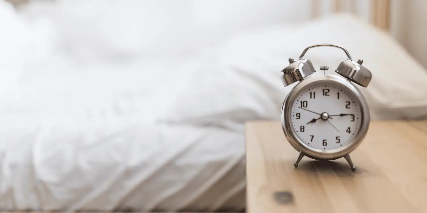 An alarm clock displaying 7:00 am, sitting on a wooden nightstand next to a bed, ready to start the day.