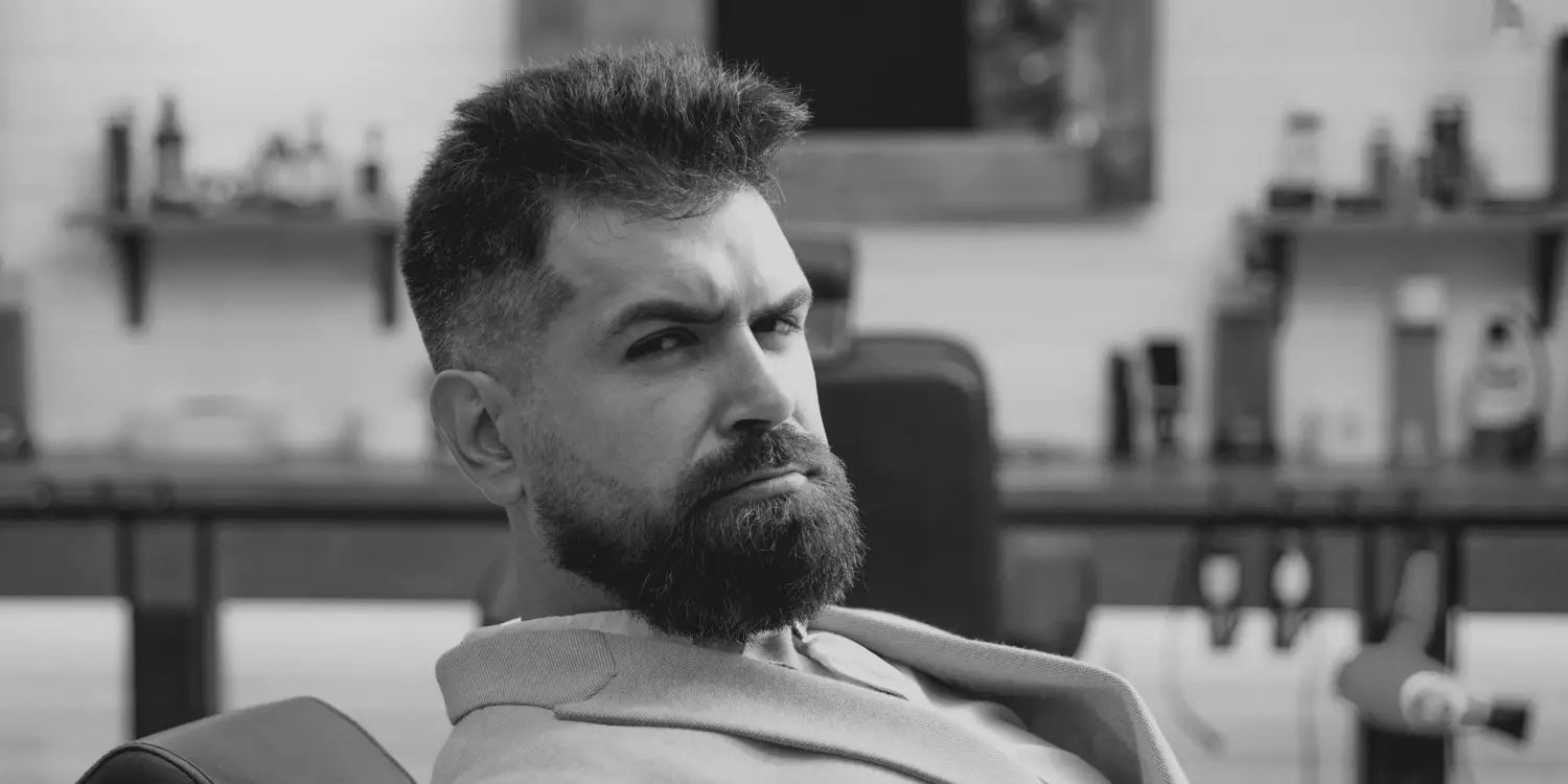 Bearded man smiling in satisfaction as he admires his freshly groomed, full beard in the mirror, while sitting in the barber's chair at a professional barbershop.