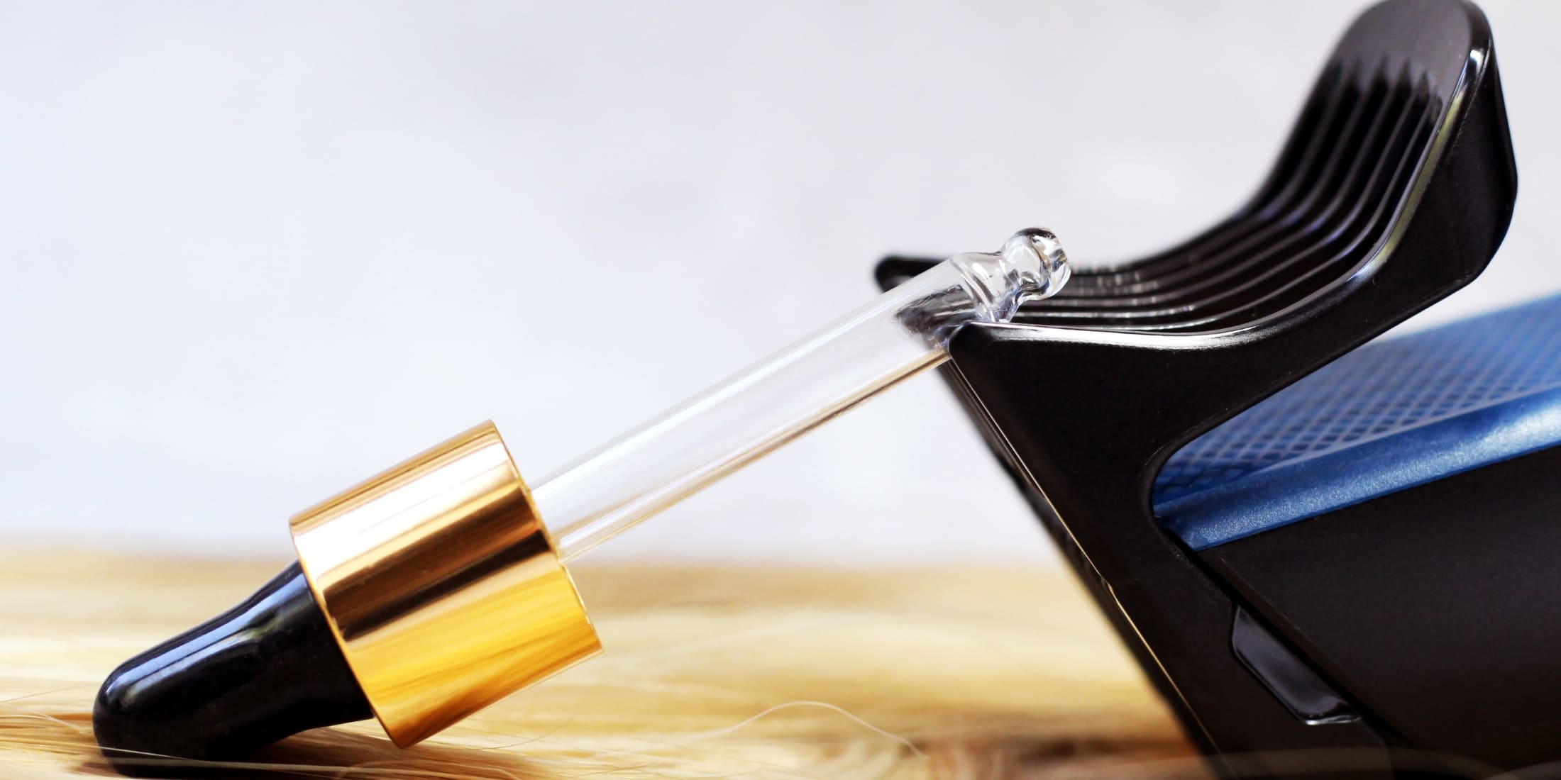A dropper bottle of clear lubricant, an hair clipper oil alternative to traditional hair clipper oil, resting on its side against a modern black and blue hair clipper, surrounded by strands of light-colored hair on a wooden surface, illustrating hair maintenance and grooming tools.