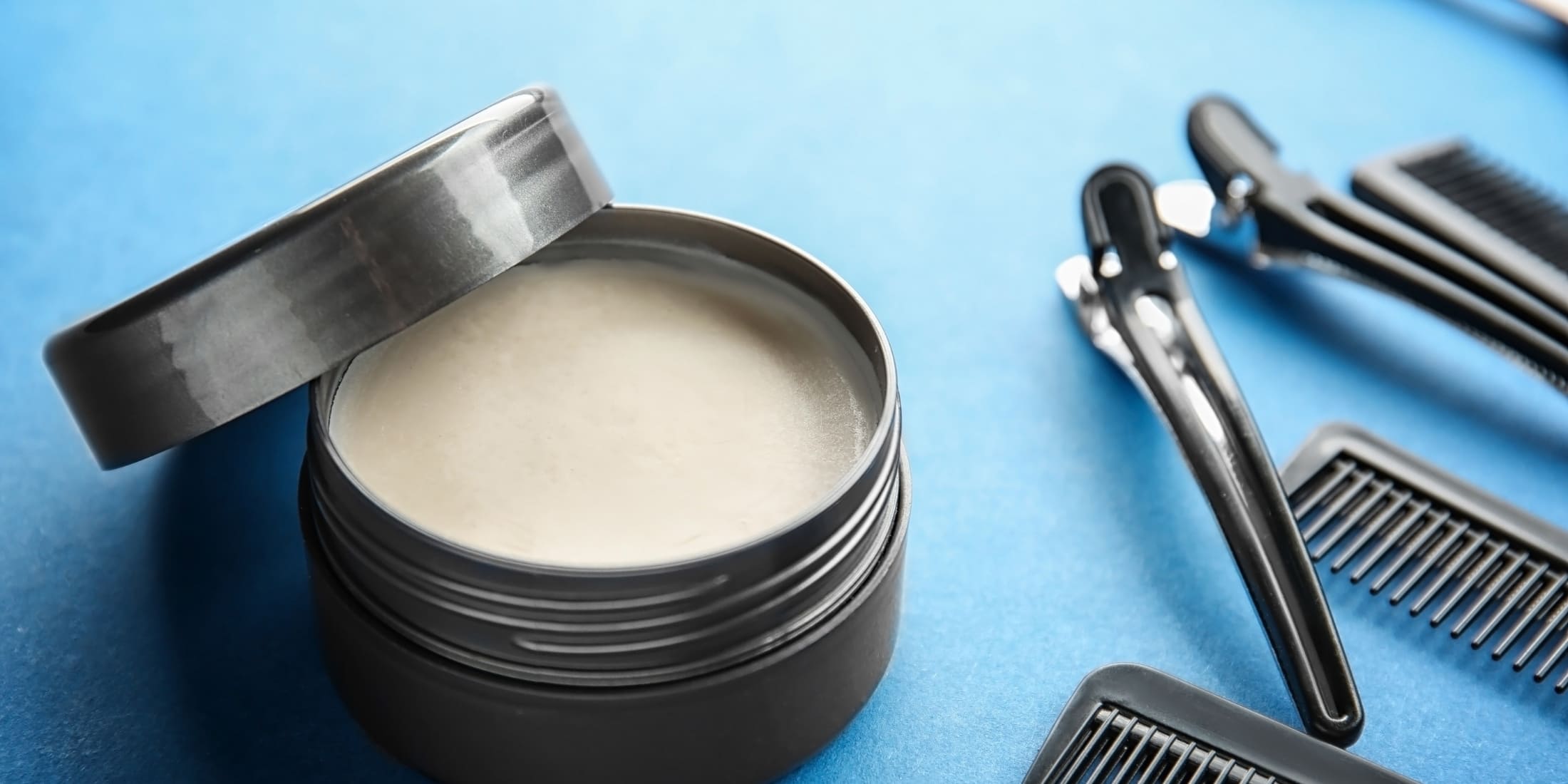 Open container of light-colored hair pomade with various black combs arranged on a vibrant blue background, showcasing essential tools for styling with pomade hairstyles.