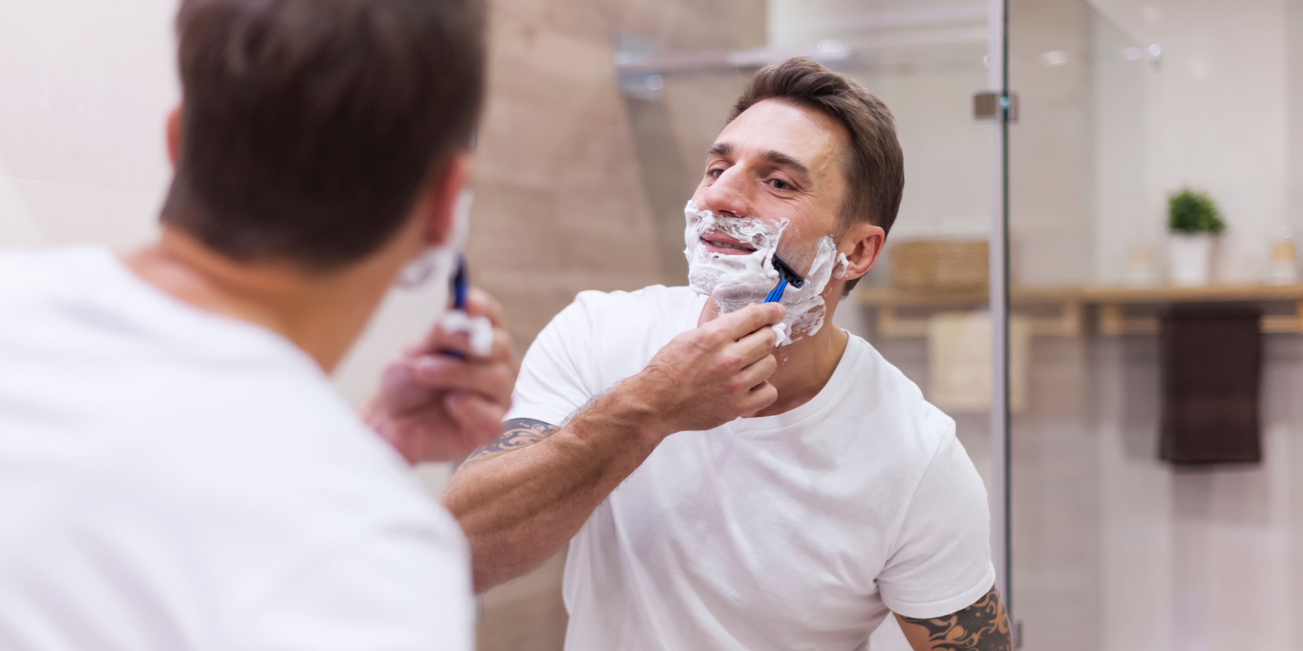 A man shaving his beard closely in front of a bathroom mirror. He is wearing a white t-shirt and has his face covered in shaving cream, focusing on achieving a smooth shave with a razor. The modern bathroom setting and his concentrated expression highlight the importance of proper shaving technique.