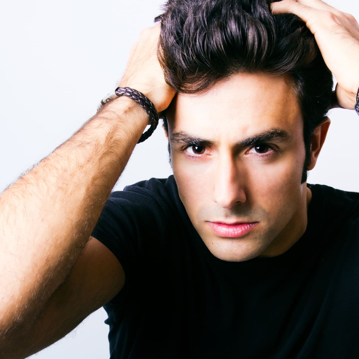 A young man with dark, thick hair is styling his hair with both hands, creating a voluminous look. He has a focused expression on his face and is wearing a black t-shirt, with a braided bracelet on his wrist. The background is plain and light, emphasizing the man's hair and grooming effort.