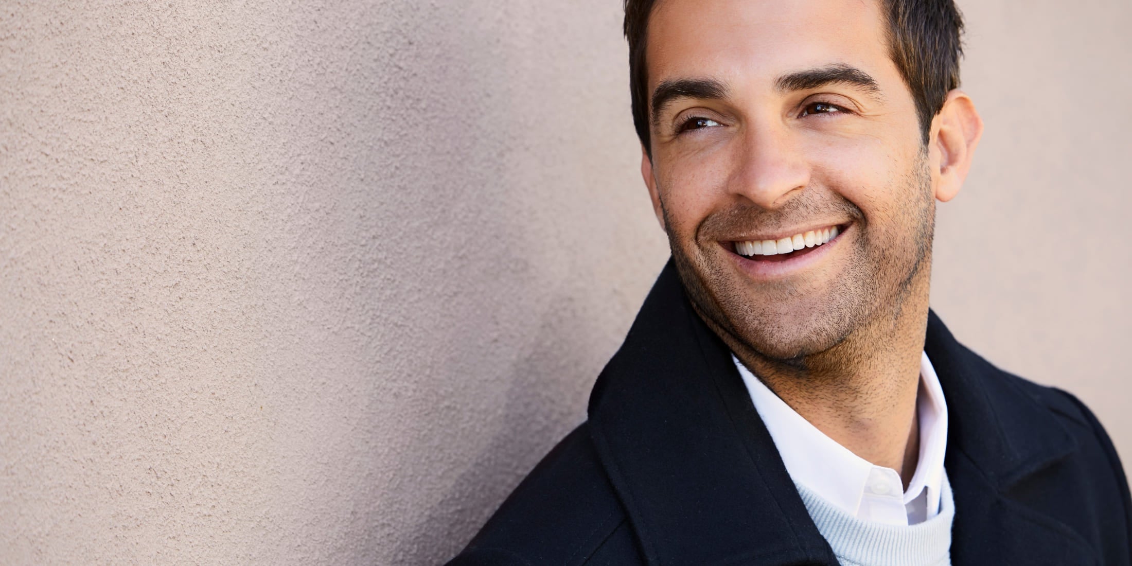 Man smiling with perfect 2-day stubble, wearing a dark blazer over a white shirt against a light-colored wall.