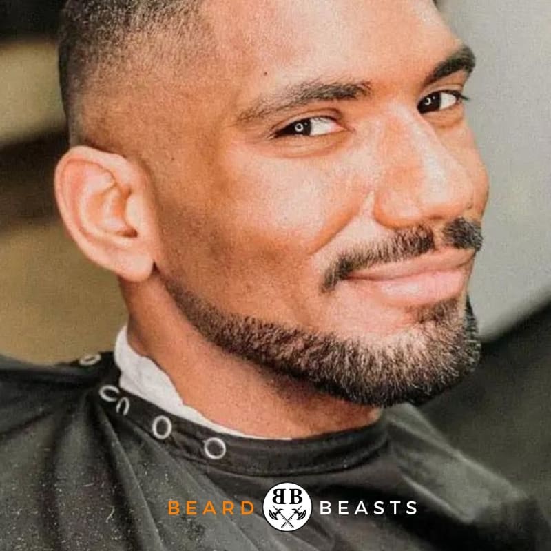 Confident man sporting a neatly trimmed Balbo beard, with a close-cut hairstyle, giving a subtle smile in a barber's cape, featuring the 'Beard Beasts' logo at the bottom.