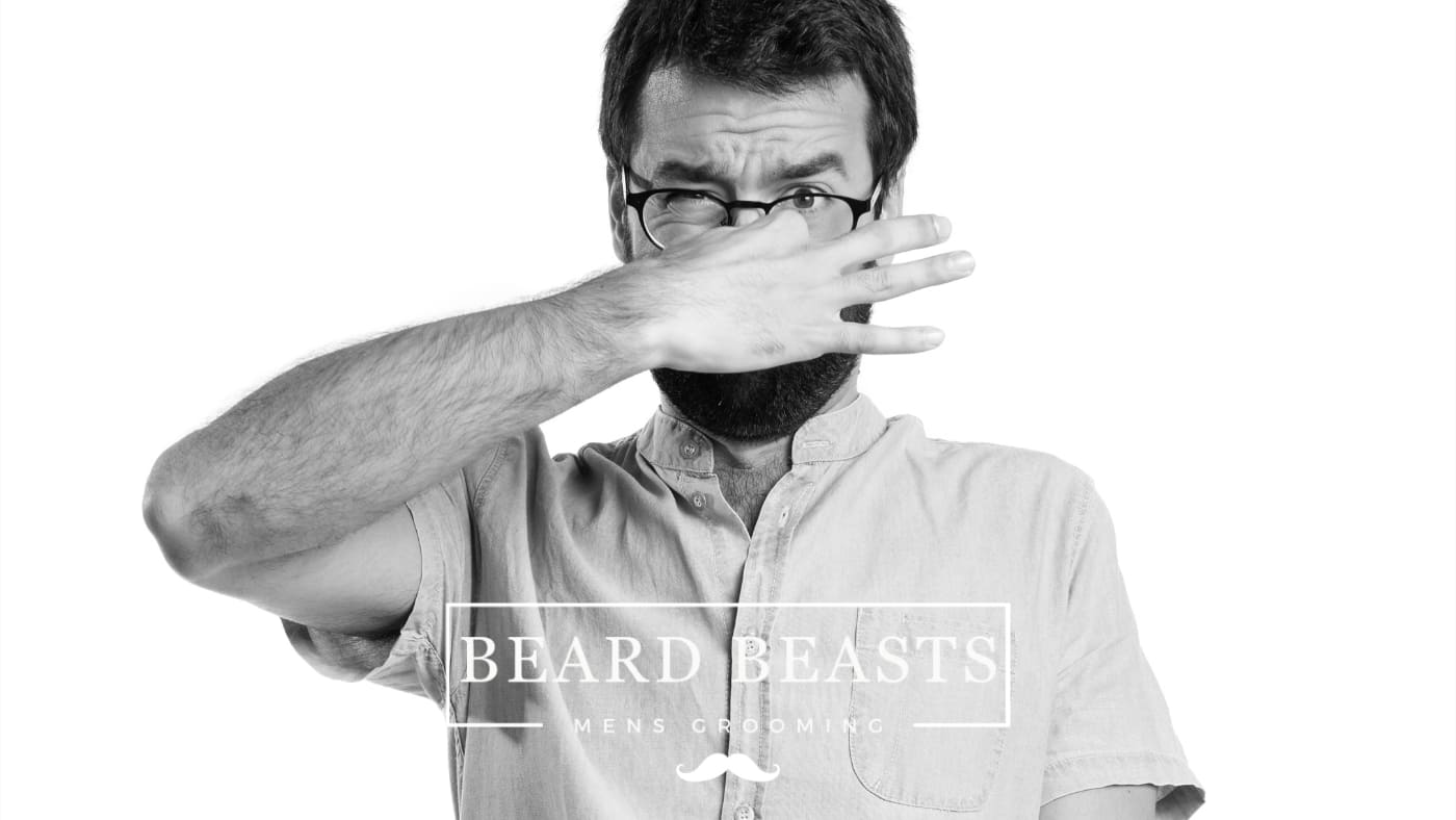 A monochrome image of a man in a casual shirt covering his nose with his hand, appearing to react to a bad smell, likely from his beard,