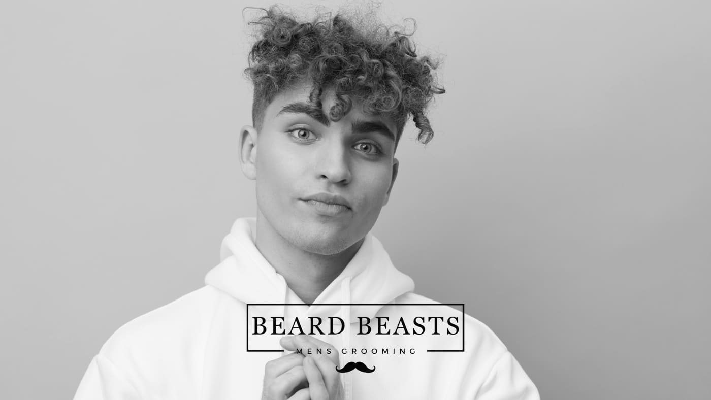 A young man with curly hair wearing a white hoodie, posing with a thoughtful expression against a gray background, with the text "Beard Beasts Men's Grooming" indicating a focus on male grooming and styling, related to 'how to get curly hair for men'.