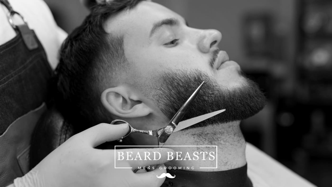A client reclines in a barber chair while a barber with precise scissors shapes his medium beard style, illustrating expert men's grooming techniques