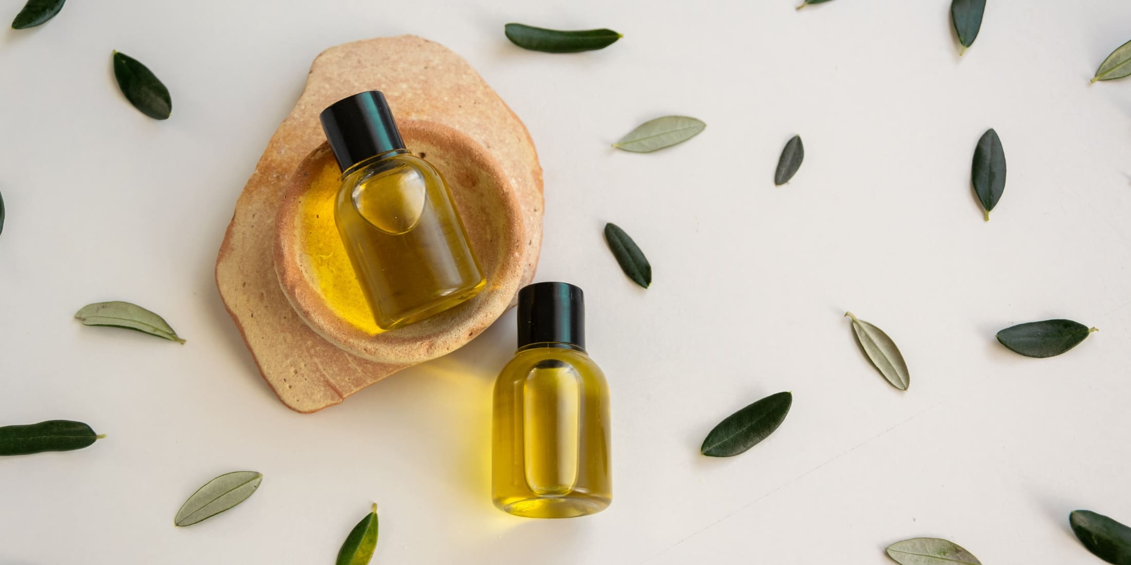 Two bottles of natural oil, potential hair clipper oil alternatives, placed on a stone dish amidst scattered green leaves on a white background, embodying eco-friendly grooming options.