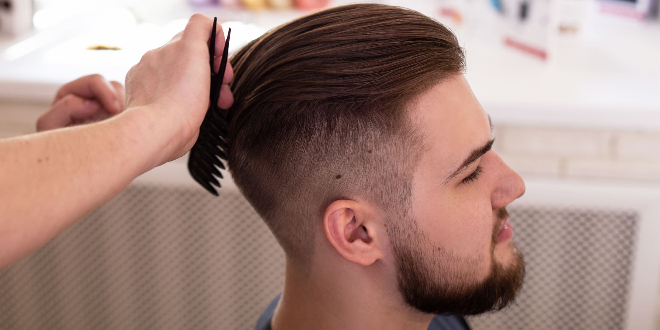 Barber styling a man's hair with an undercut fade, highlighting the undercut vs taper contrast with long, slicked-back top and short, faded sides.