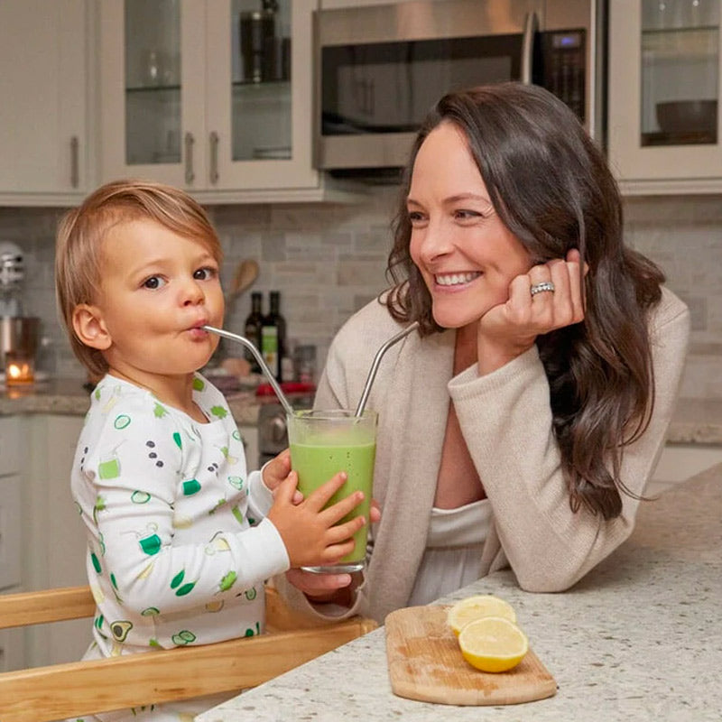 A toddler sips a healthy green smoothie with the help of his smiling mother.