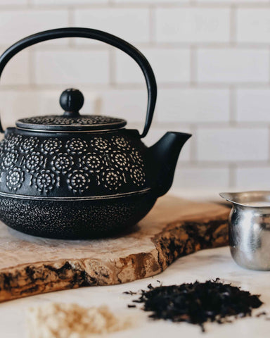 Black textured teapot sits on a wooden board as there is loose leaf tea sitting in the foreground