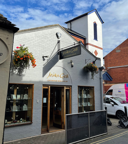 The shop front of Mike Gell Jewellery in Hereford. It shows a white-painted building with angled eaves and a small tower, hanging baskets and a display of jewellery in the front windows.