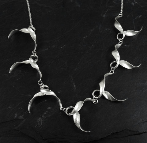 A silver necklace of seven twisted, looping forms like wings in flight joined by a loop where the bird's body would be