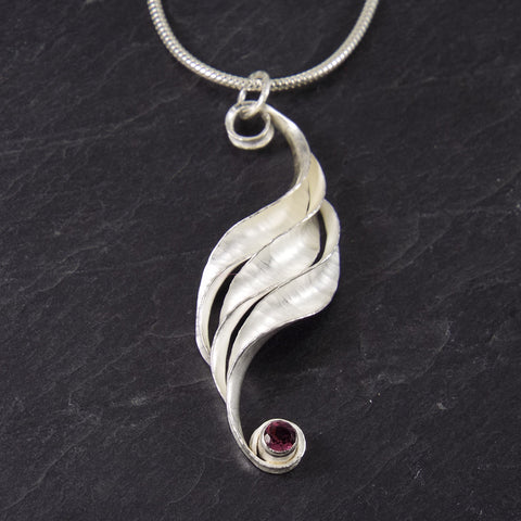 A silver pendant, handmade, consisting of three curled and twisted units which form a progression, with a garnet at the bottom end.