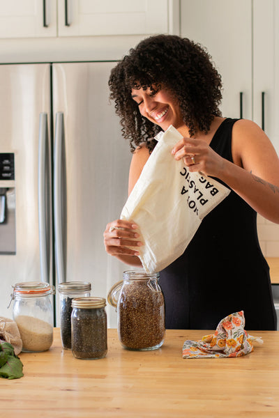 A Black, female model stands in a kitchen pouring contents out of a Market Bags "Not a Plastic Bag" reusable produce bag into a jar.
