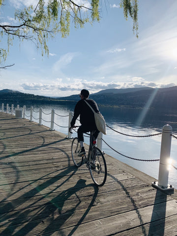 a young man riding a bike down a board walk along the lake with a bag slung over his shoulder
