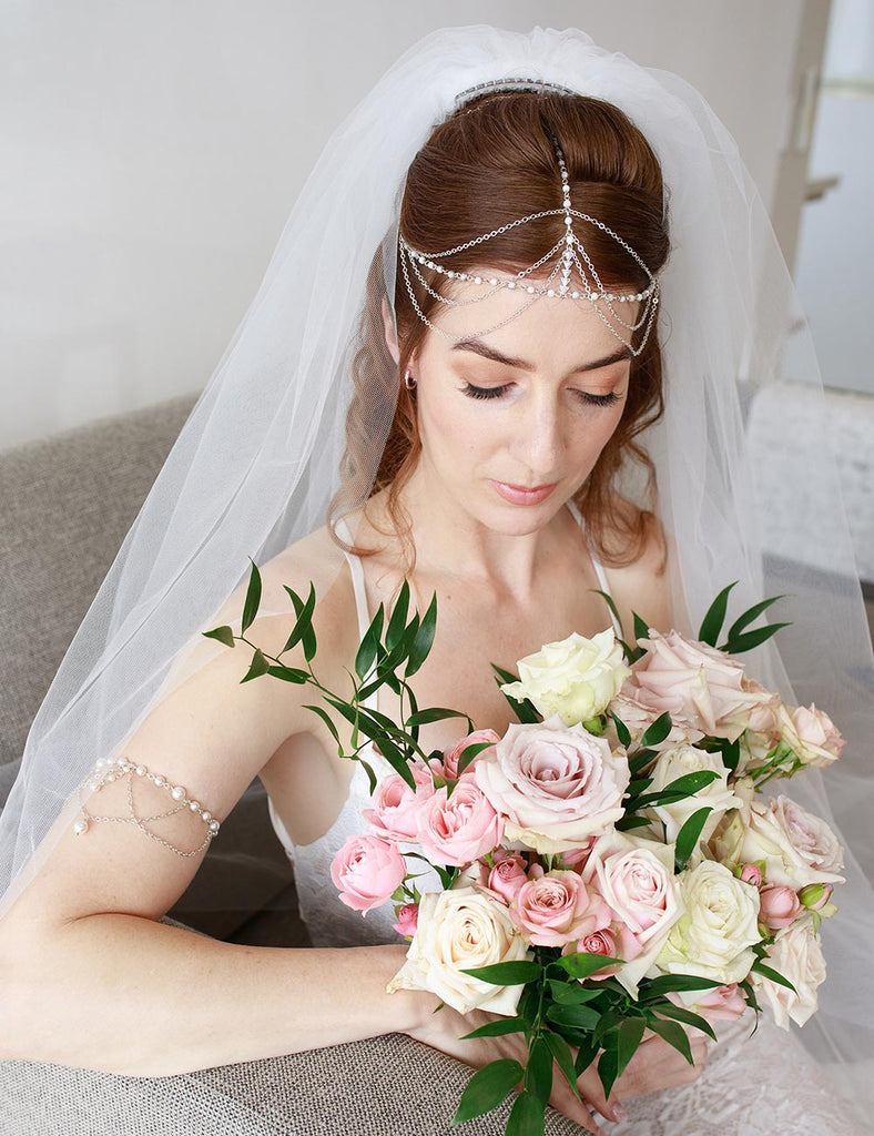 Bride Frances sits on couch looking down into flower bouquet wearing custom head chain with veil and arm bracelet