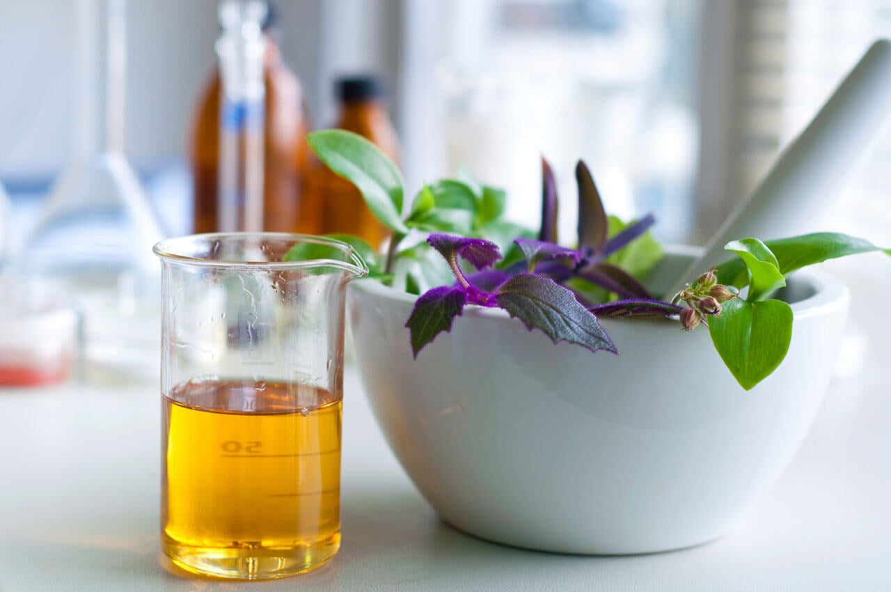 Essential oil in beaker next to pestle with herbs.