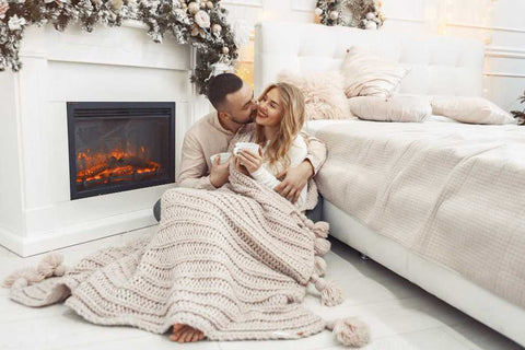 A man and a woman holding mugs, cuddling in front of the fireplace.