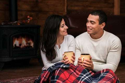 Man and woman smiling sitting next to eachother and holding mugs in front of the fireplace.
