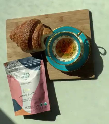 Maisha chai tea in a tea cup, next to a package of chai and a half-eaten croissant.