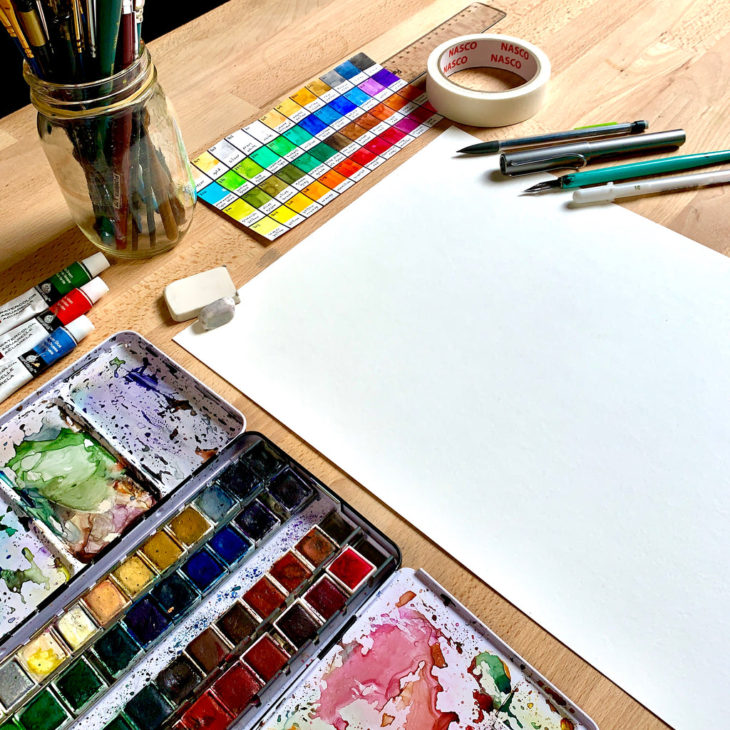 Close up shot of watercolor painting materials on a wooden desk surrounding blank paper