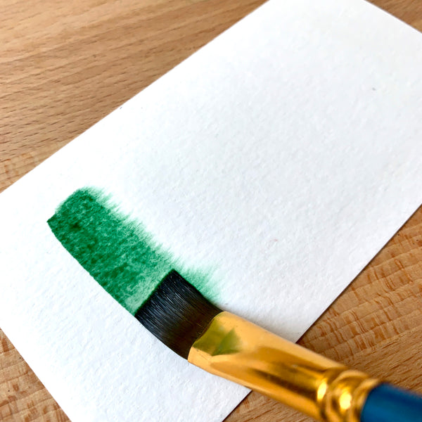 Close up of a paint brush filled with dark green paint on watercolour paper