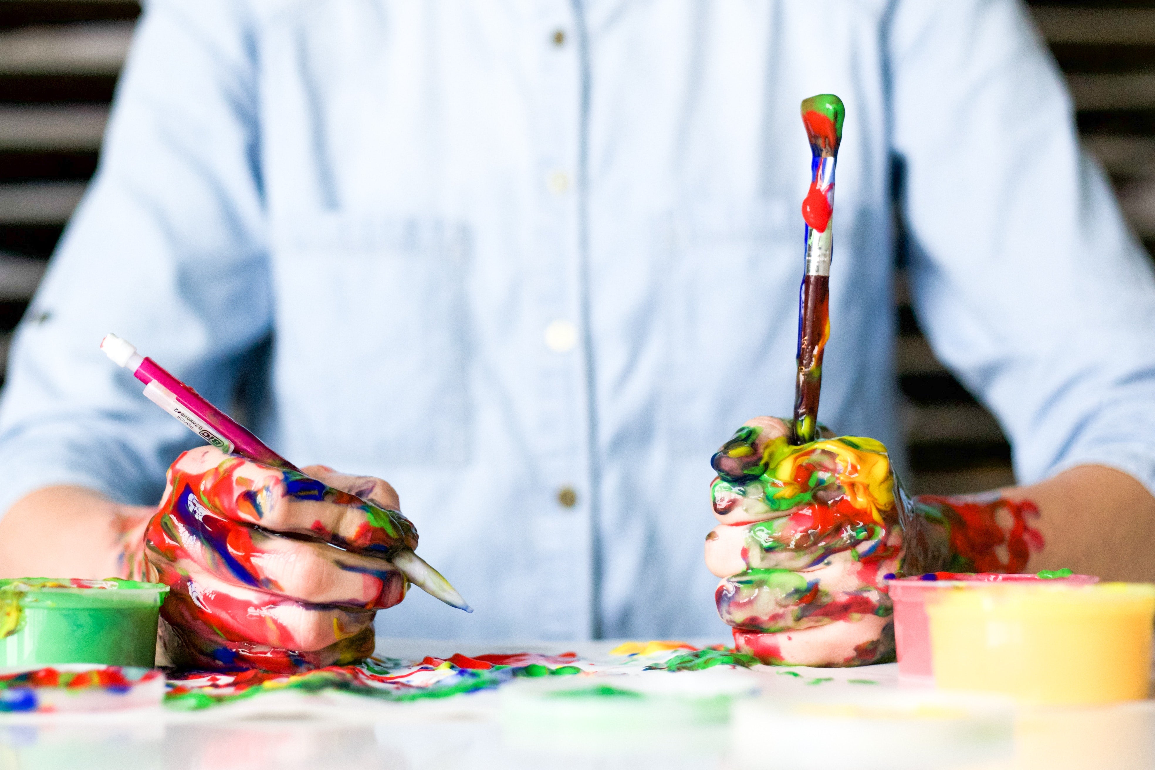 Man in blue shirt holding paintbrushes in both hands with paint everywhere 