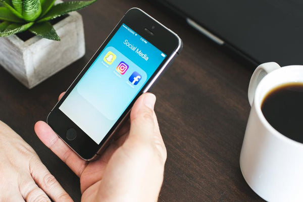 Close up shot of a hand holding a phone with social media icons present next to a cup of coffee on a desk