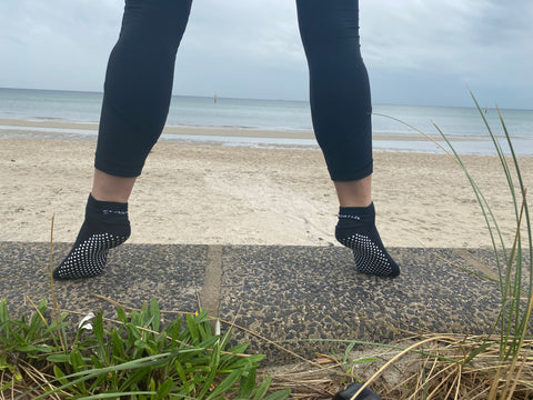 Image of Stealth Movement Grip Socks worn on a concrete surface with a beach in the background. The model is standing on her toes, showcasing the white grips underneath.