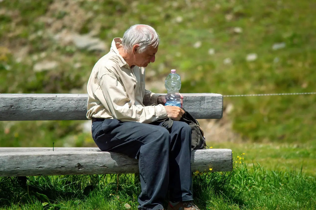 Elderly walker having a rest on a bench whilst drinking water to rehydrate during hot weather