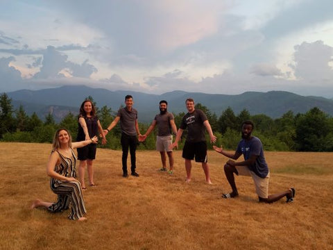 6 members of the Compt team stand and kneel in a field with clouds overhead in front of a beautiful forested mountain range.