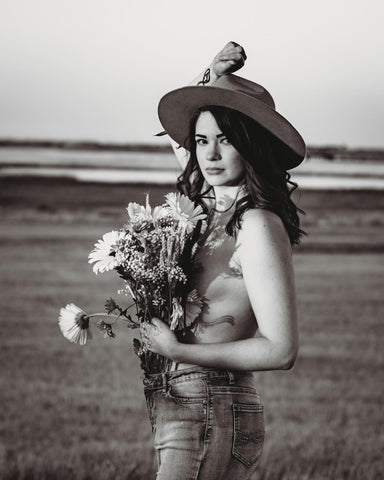 Image of Kelsey Fay, standing in a field, wearing a hat, and holding wildflowers