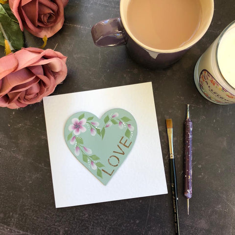 White card decorated with blossom flowers and stencilled lettering reading ‘love’ surrounded by a candle, mug of tea and flowers  