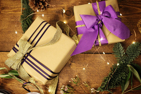 two presents are wrapped in brown paper with gold and purple trimmings. It is surrounded by festive odds and ends like pine cones, ribbon, pine sprigs and tiny fairy lights.